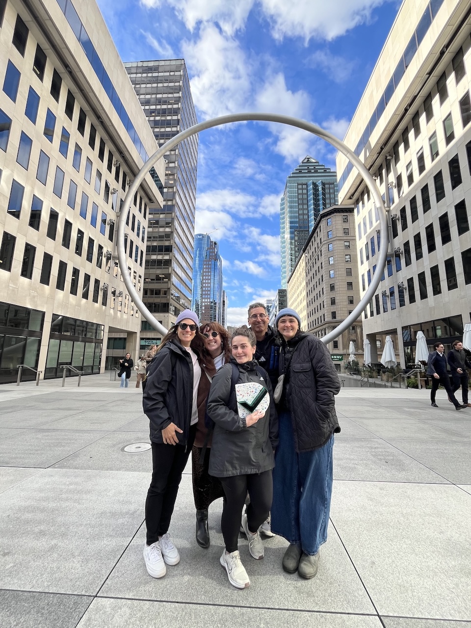 Cinq personnes d'Overall pose ensemble dans une rue de la ville, encadré par une architecture moderne et une sculpture circulaire, sous un ciel nuageux.