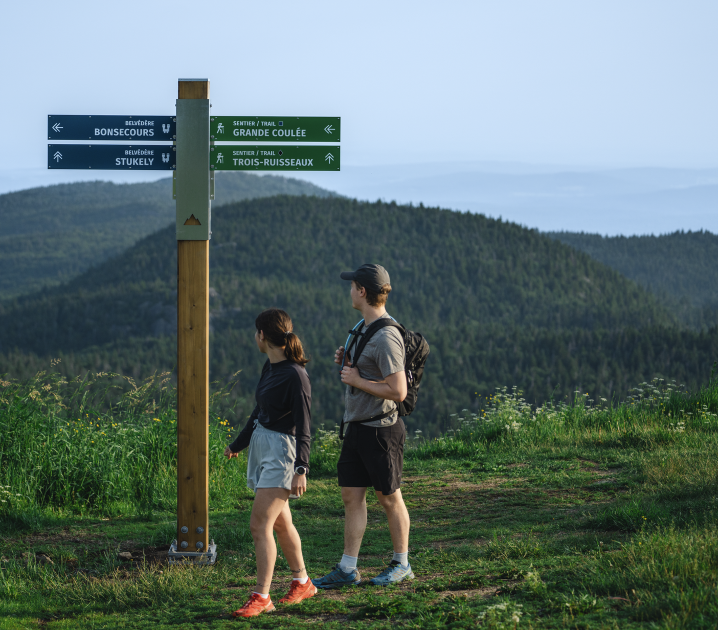 Deux randonneurs consultent un panneau de sentiers en montagne, avec vue panoramique sur les collines boisées.
