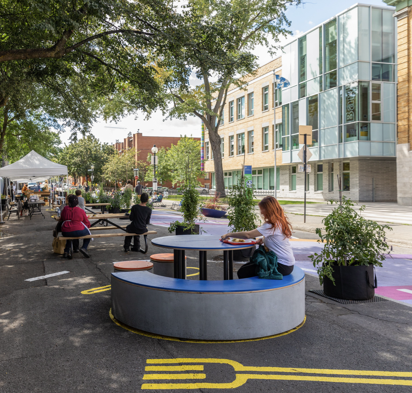 Un espace de restauration en plein air au Bleu Tomate, avec des tables rondes, de la verdure et des gens qui savourent leur repas dans un cadre urbain animé.
