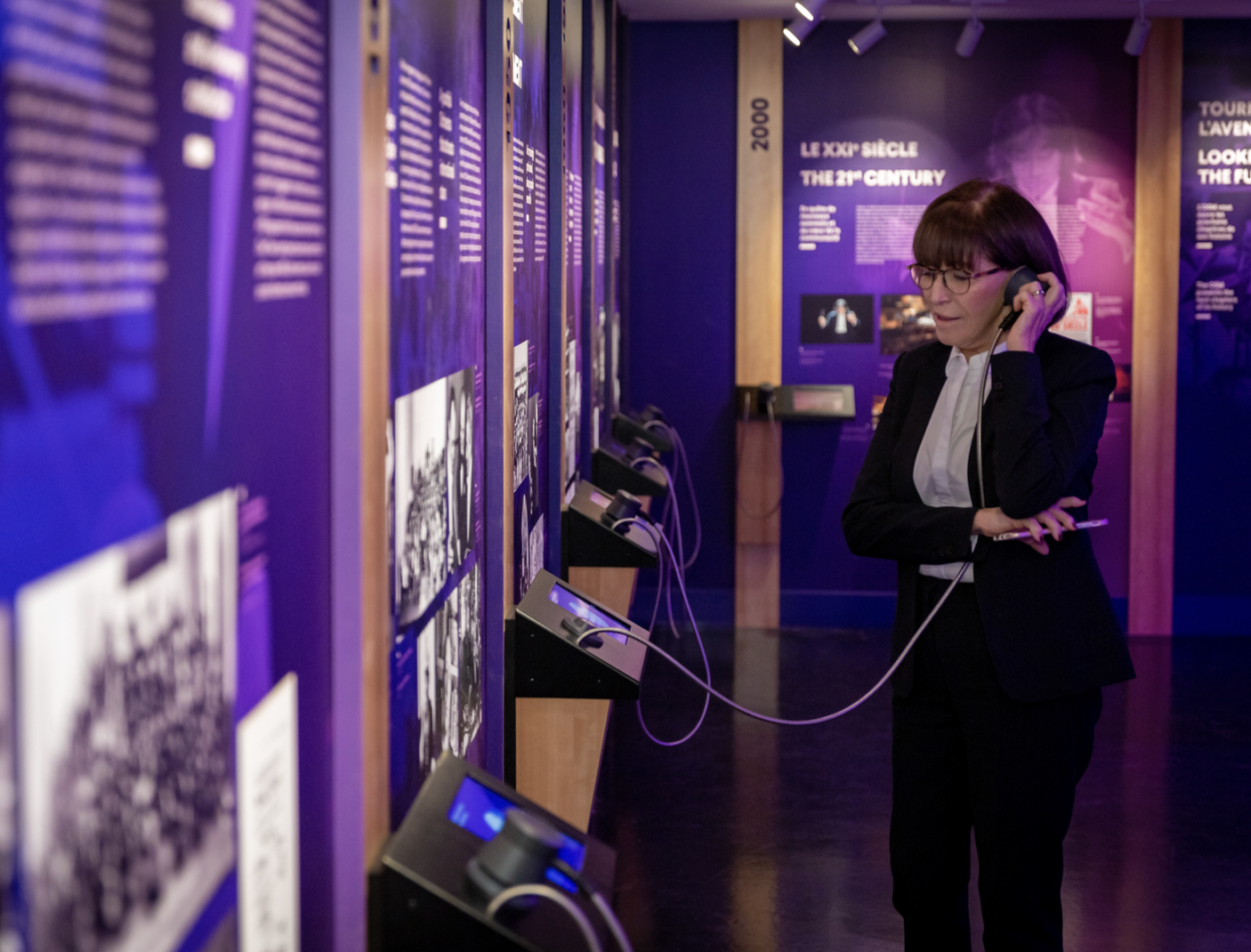 Un visiteur en tenue formelle écoute un audioguide devant les panneaux d'information d'une exposition dans un musée.