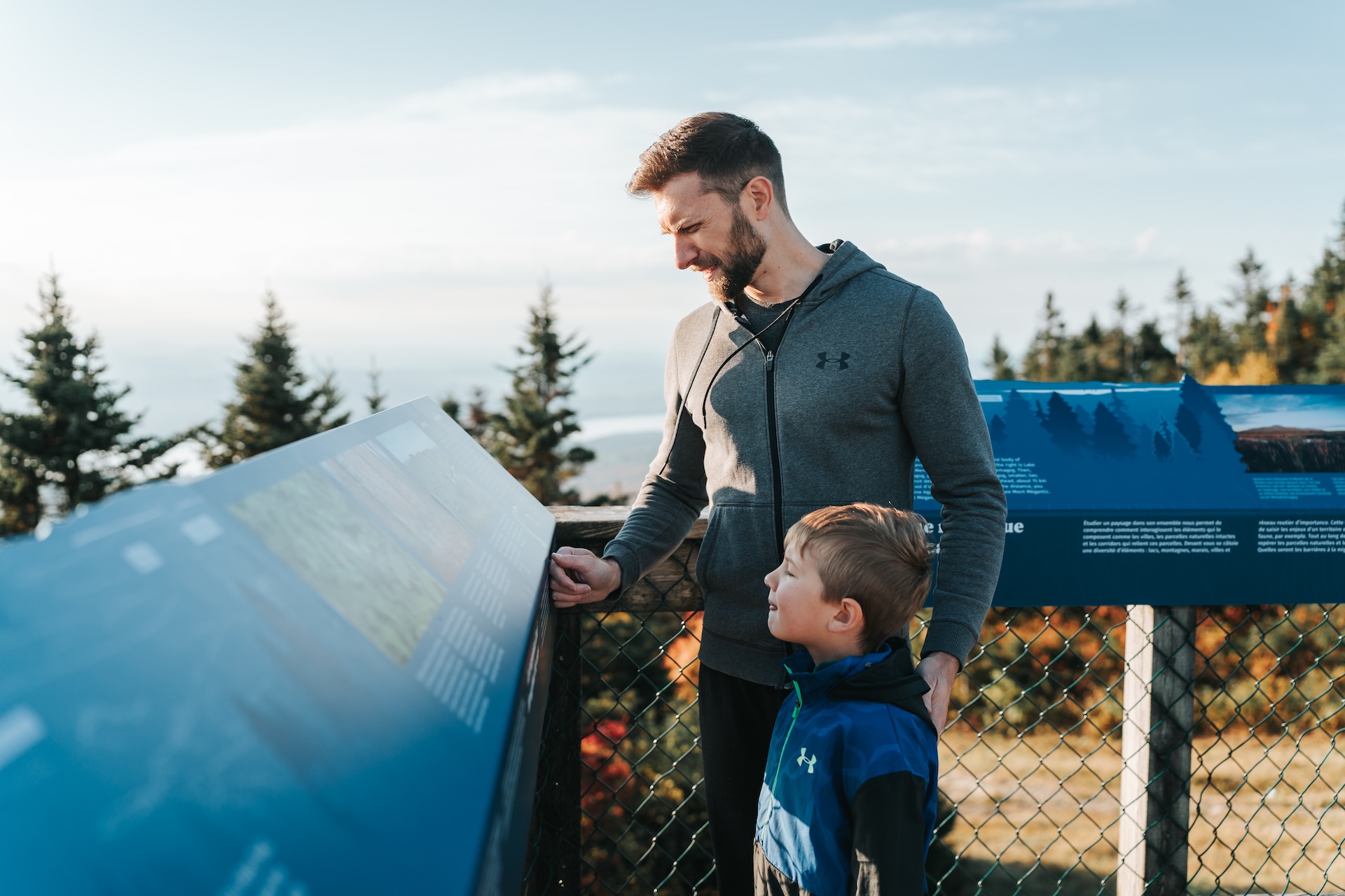 Père et fils observent une plaque d'information en extérieur, dans un cadre naturel boisé.
