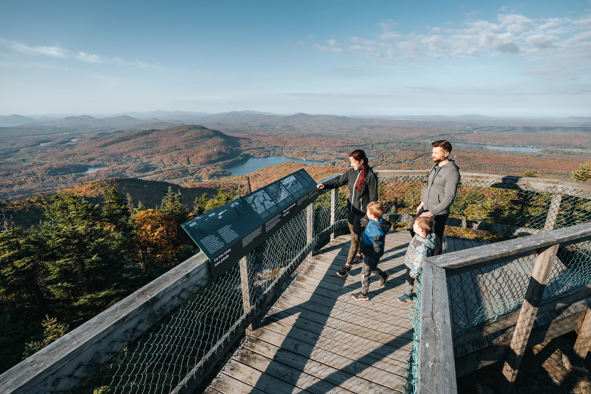 Famille admirant un panorama automnal depuis une passerelle en bois avec panneau d'information.