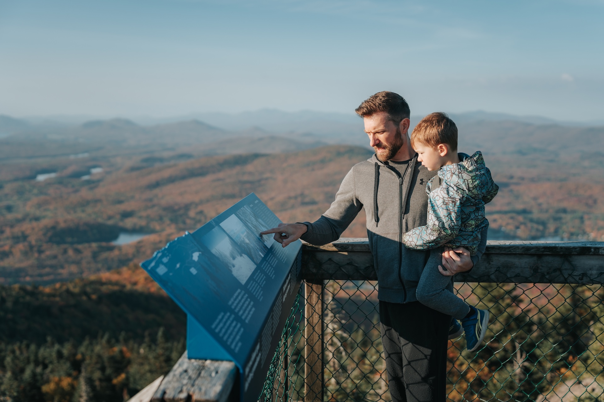 Père et enfant observent un panneau d'information sur un balcon de montagne en automne.