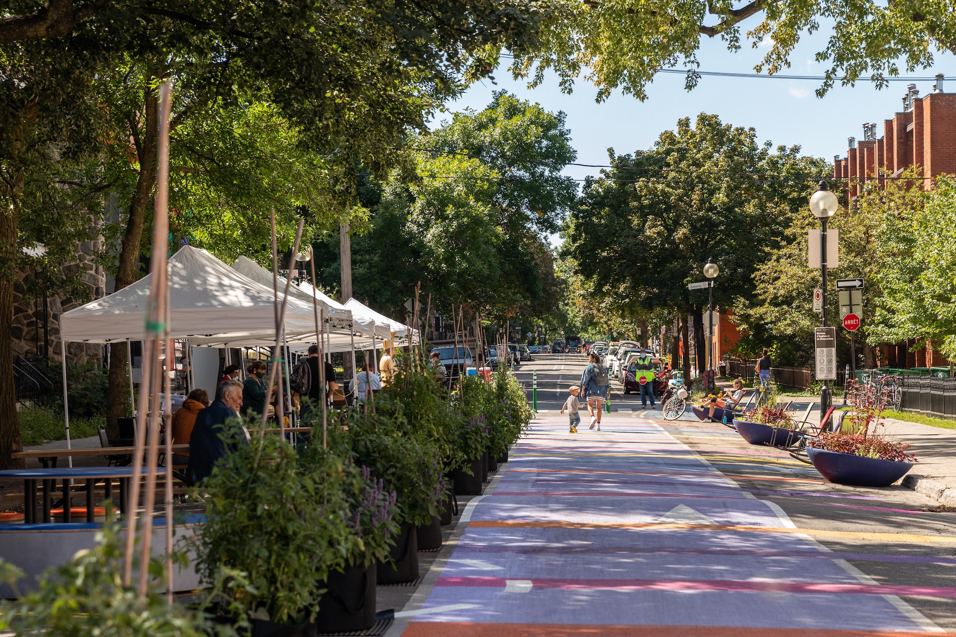 Rue transformée en espace public avec terrasse de café, arbres et piétons, projet urbain de Studio Overall.