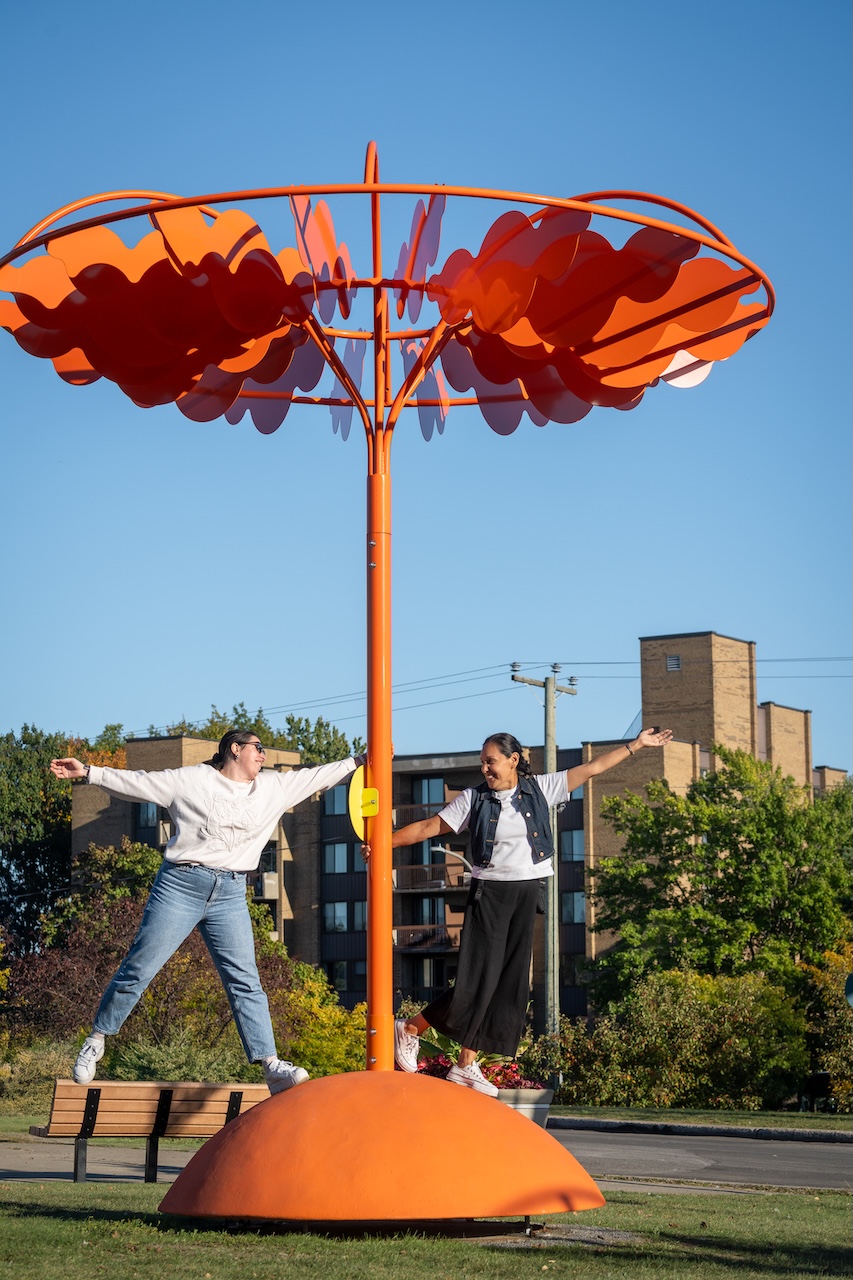Deux personnes posent autour d'une sculpture orange en forme de fleur dans un parc urbain.