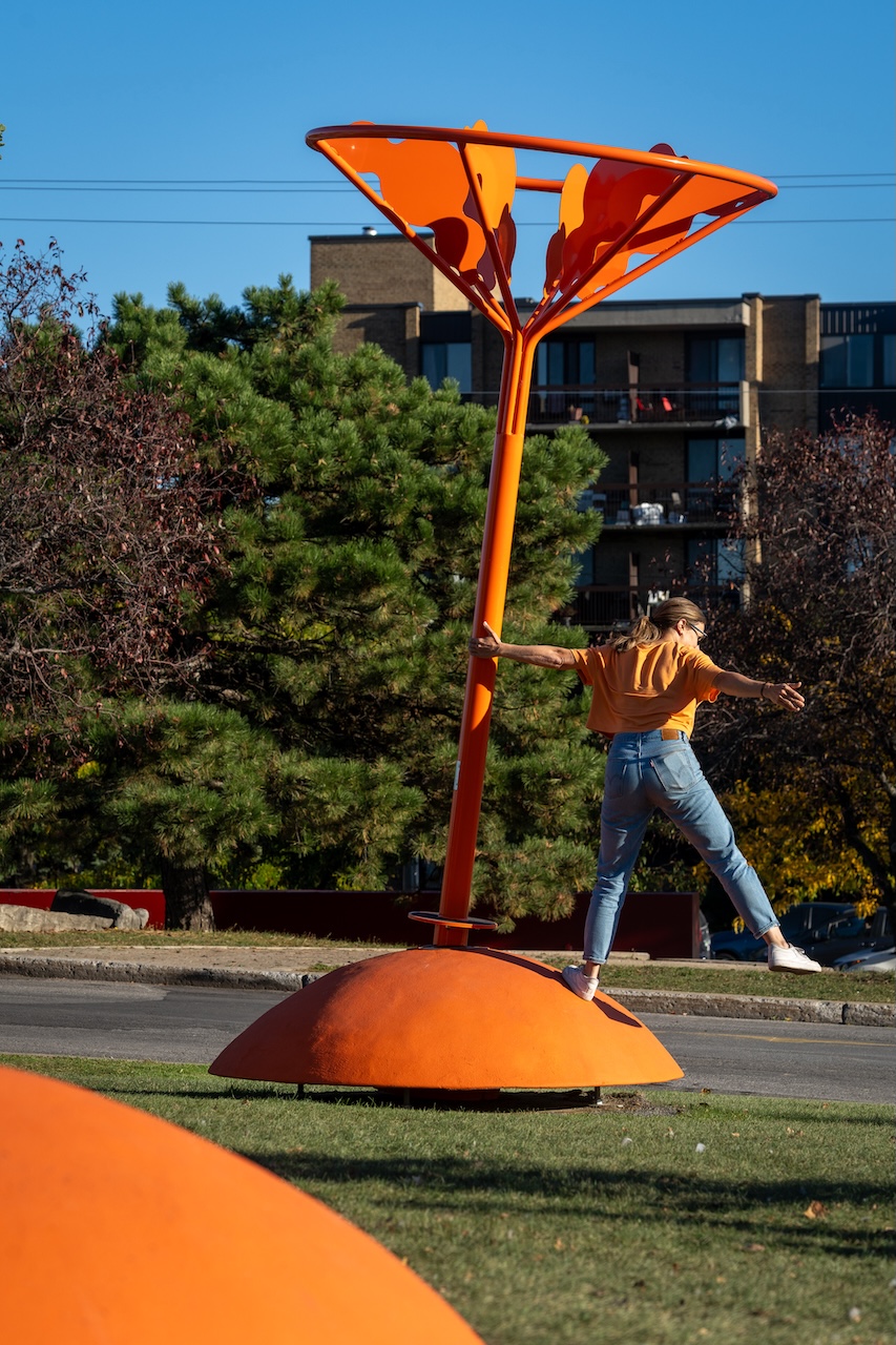 Installation artistique orange en plein air, avec une personne s'y appuyant dans un parc urbain.