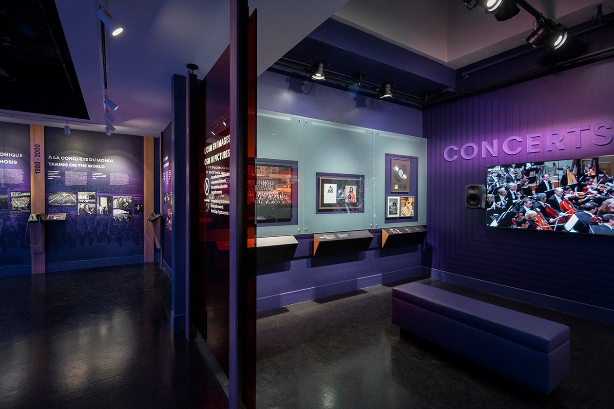 Exposition muséale immersive avec écrans, vitrines et sièges pour visiteurs.