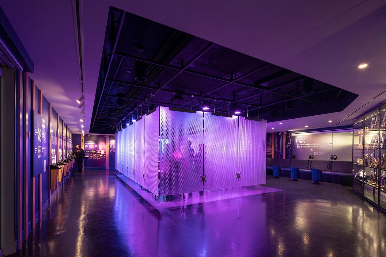 Installation muséale lumineuse avec cloisons perforées, ambiance violet, espace d'exposition moderne.