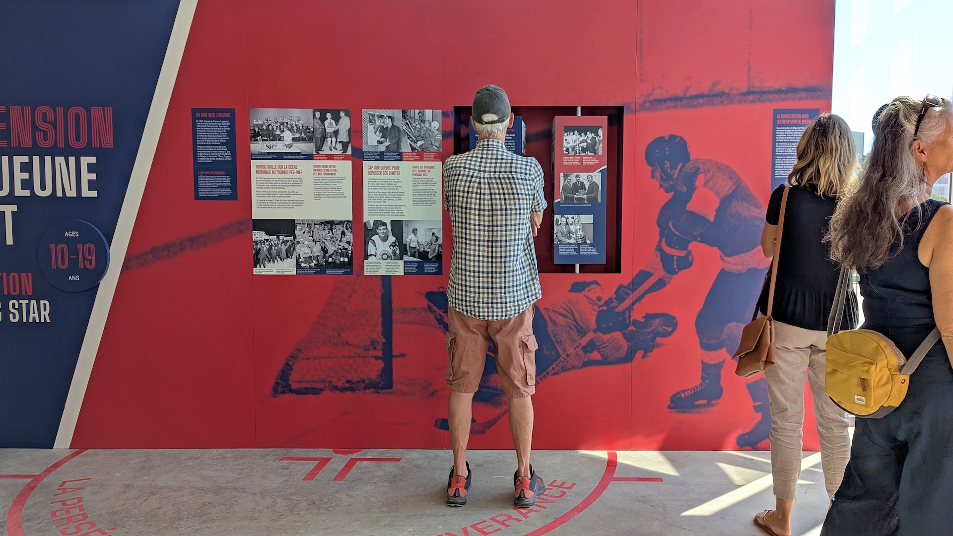 Visite d'exposition sur l'histoire du hockey, avec visiteurs devant un mur rouge orné de photos et de silhouettes de joueurs.