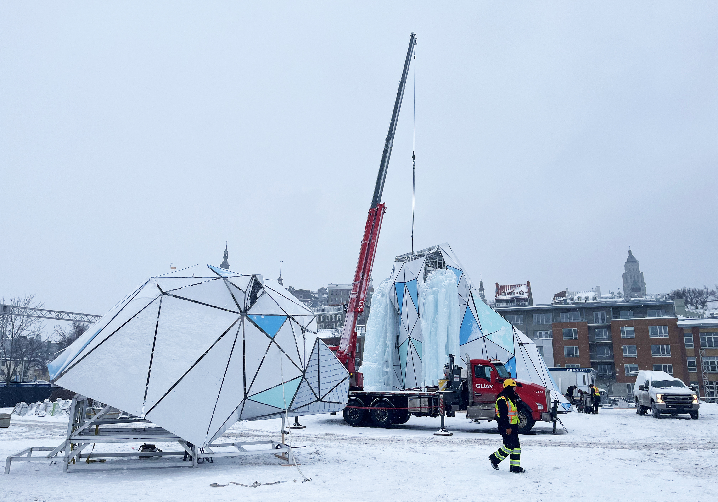 Installation artistique enneigée en cours de montage avec une grue, dans un cadre urbain hivernal.
