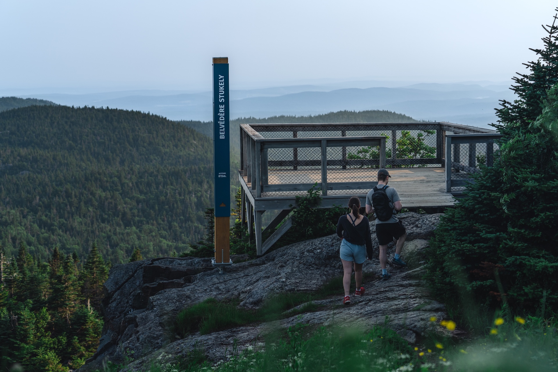 Vue panoramique depuis un observatoire en bois, avec deux personnes marchant sur un sentier rocheux.