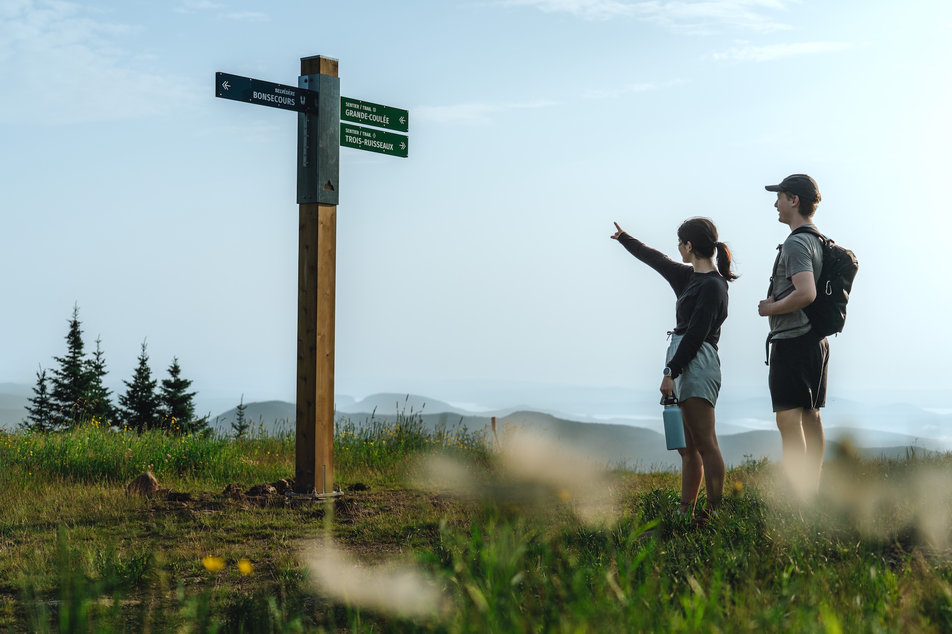 Deux personnes observent un panneau de sentiers en montagne, avec vue panoramique.