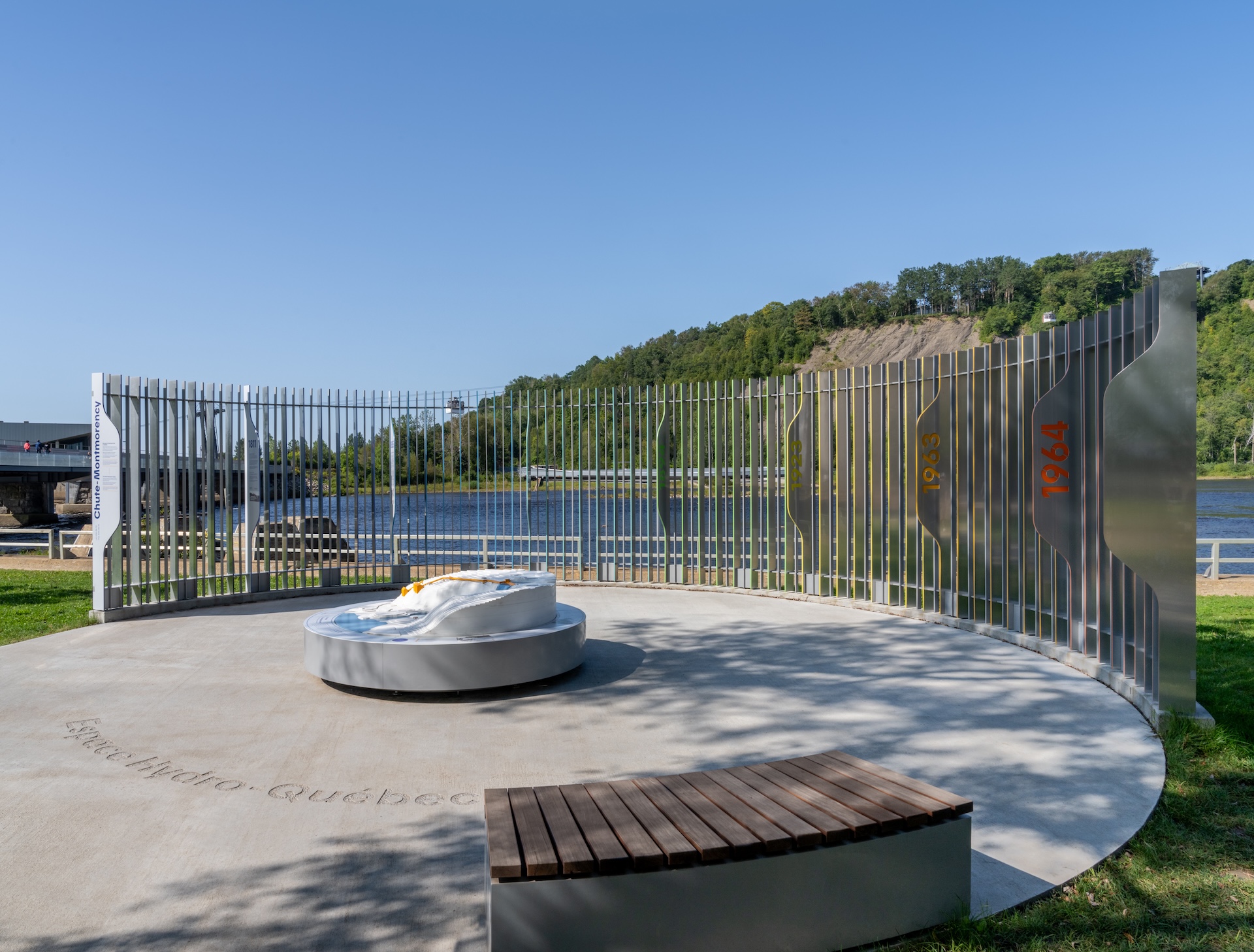 Installation contemporaine en bord de rivière, avec mémorial en métal et sculpture circulaire.