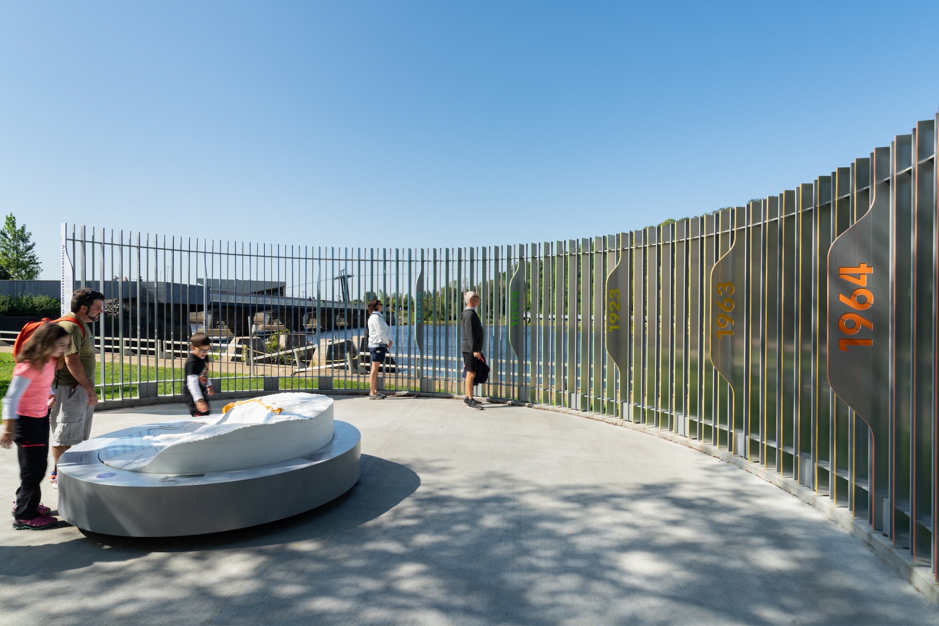 Installation contemporaine avec visiteurs, mémorial en métal avec dates, espace aquatique en arrière-plan.